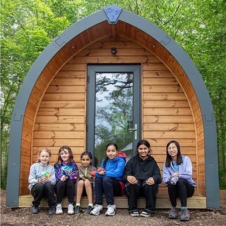 school group sitting by a wooden log cabin in a forest area